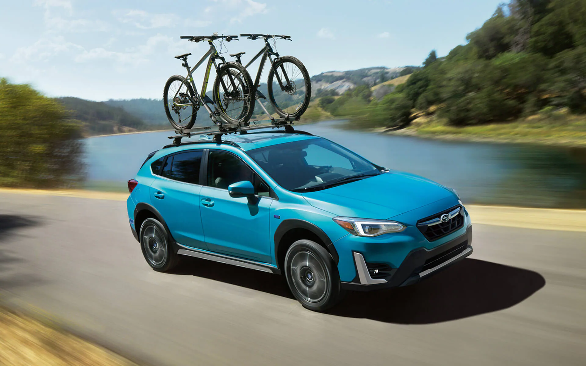 A blue Crosstrek Hybrid with two bicycles on its roof rack driving beside a river